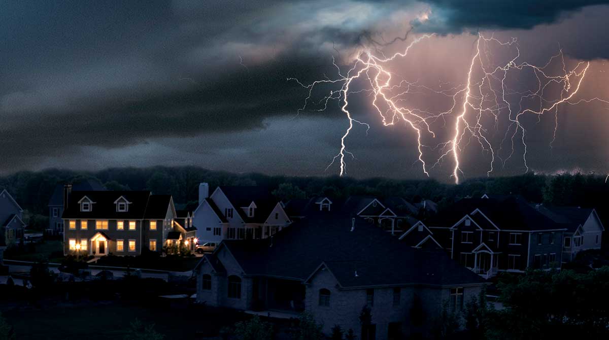 Lightning over a dark neighborhood power outage while a single home has power and lights supplied by a generator while surrounding homes remain dark.